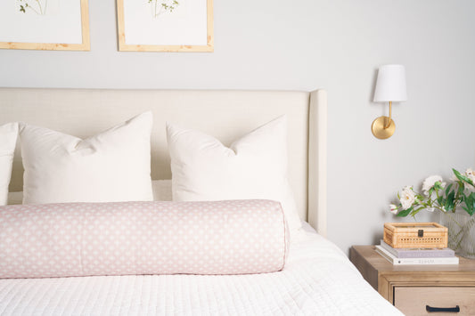 Neatly made bed with white bedding and pink decorative pillow, nightstand with books and flowers, framed pictures on wall.